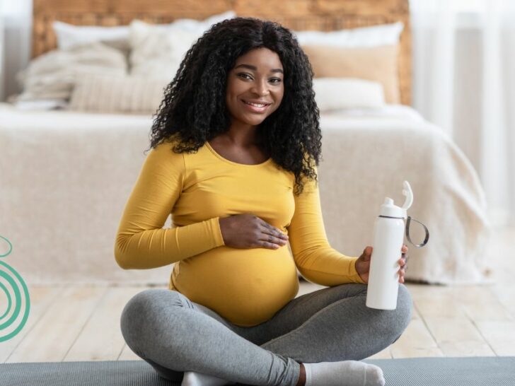 black pregnant woman with yellow shirt sitting on a yoga mat with a water bottle